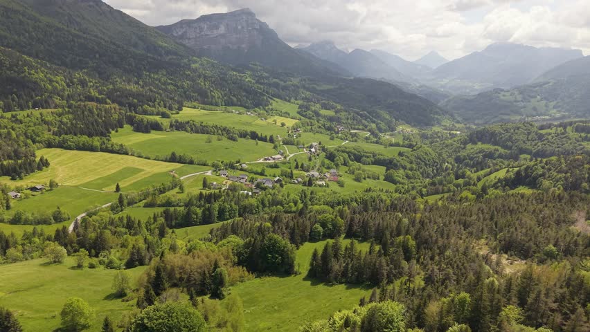 Aerial orbital drone shot of a village in the Chartreuse Valley, French Alps, France. Scenic mountain landscape with valley, alpine peaks, clouds and overcast sky, cinematic aerial view.