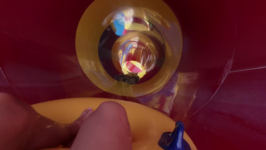 POV video of person riding colorful funnel water slide in aquapark, feet visible in inflatable tube, splashing water and bright striped walls on sunny summer day