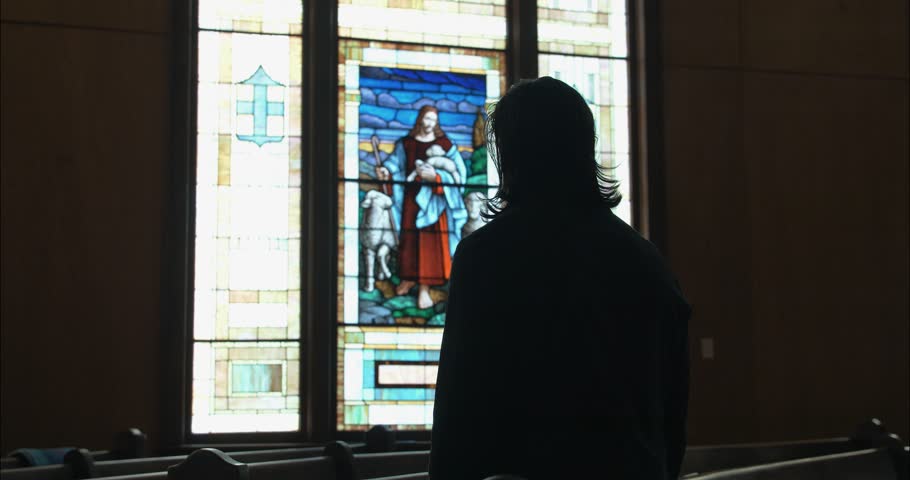 A man standing inside a church, gazing at a stained-glass window of Jesus the Good Shepherd, evoking themes of spirituality, faith, and quiet reflection.