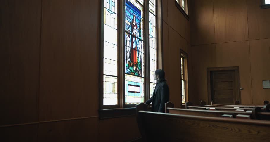 A Christian man, priest, preacher, pastor in spiritual reflection and worship while praying inside old church building, by stained glass window creating a cinematic and contemplative mood.