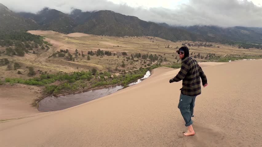 Person running down the Great Sand Dunes National Park Sangre de Cristo range Rocky Mountains spring summer raining cloudy Colorado unique magical large amounts of sand dune hills windy landscape pan