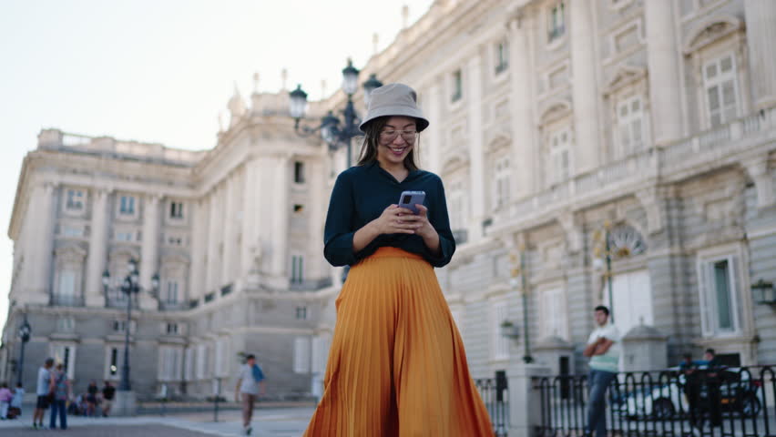 Smiling Chinese woman tourist standing in front of Royal Palace Madrid using smartphone. Symbol of sightseeing, cultural exploration, and digital travel lifestyle