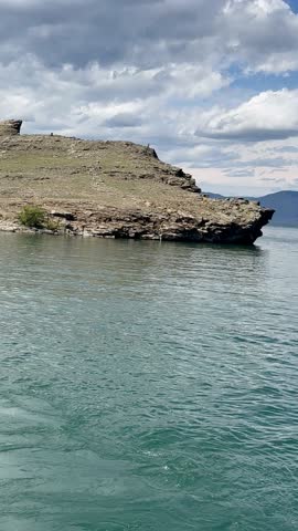 Baikal Lake on sunny day. Vertical video of ferry sailing from village of Sakhyurta past coastal rocks to Olkhon Island. Beautiful blue sky with cumulus clouds over blue water. Natural background