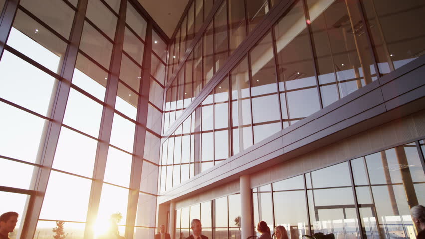 Two young businessmen who are old acquaintances, meet and shake hands in a busy modern office building at sunset. 