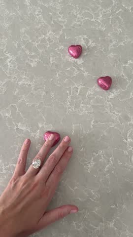 Romantic vertical video showing an oval diamond engagement ring or wedding band placed beside pink heart chocolates on a marble background symbolizing Valentines Day love.