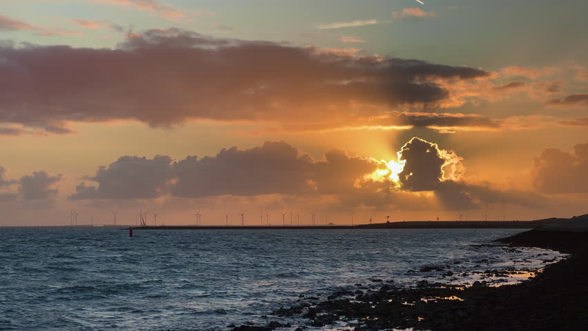 Spectacular sunset with dramatic orange and grey clouds over wind farm and sailing boat. Golden sunbeams break through storm clouds above ocean waves on rocky Zeeland coast Netherlands.