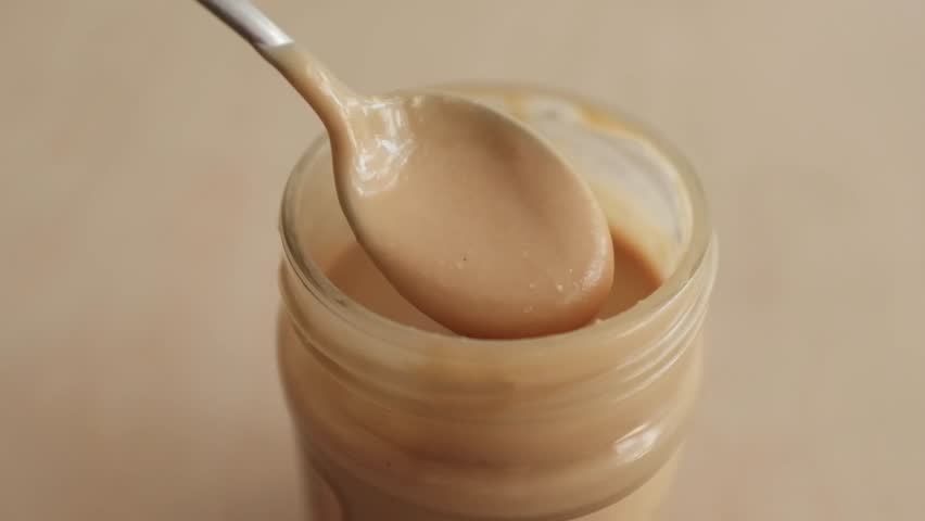 Closeup video of peanut butter in a jar. A spoon is mixing liquid peanut butter with lumpy texture.