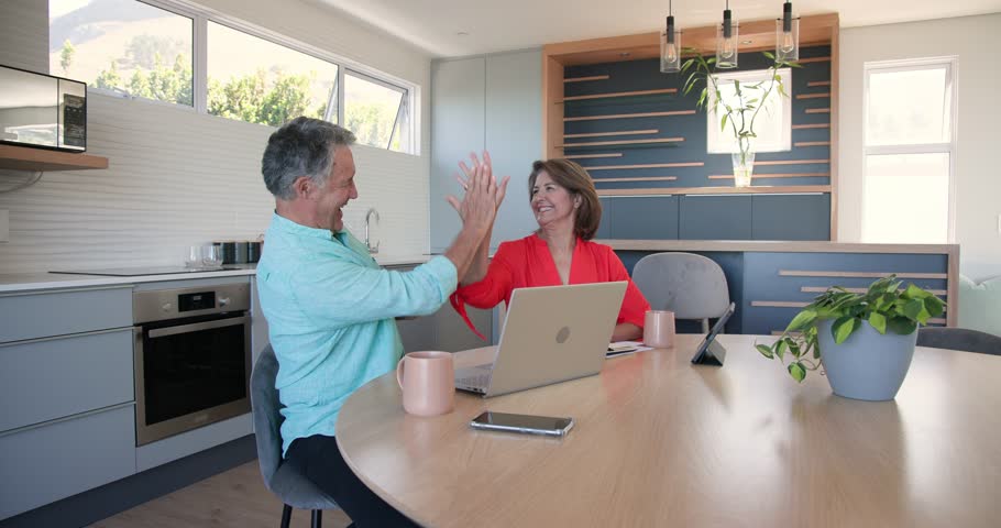 Couple celebrating win exchanging high five typing on silver laptop at dining table sharing ideas. Partnership, collaboration, communication, leisure, modern, cozy, companionship
