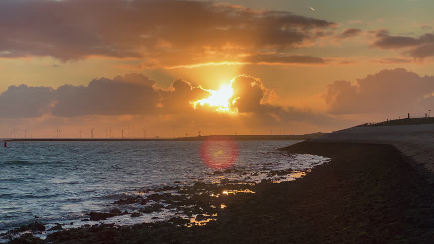 Golden sunset with brilliant sun reflection on ocean water and wind farm on horizon. Dramatic orange clouds above rocky shore with waves washing coastline in Zeeland Netherlands.