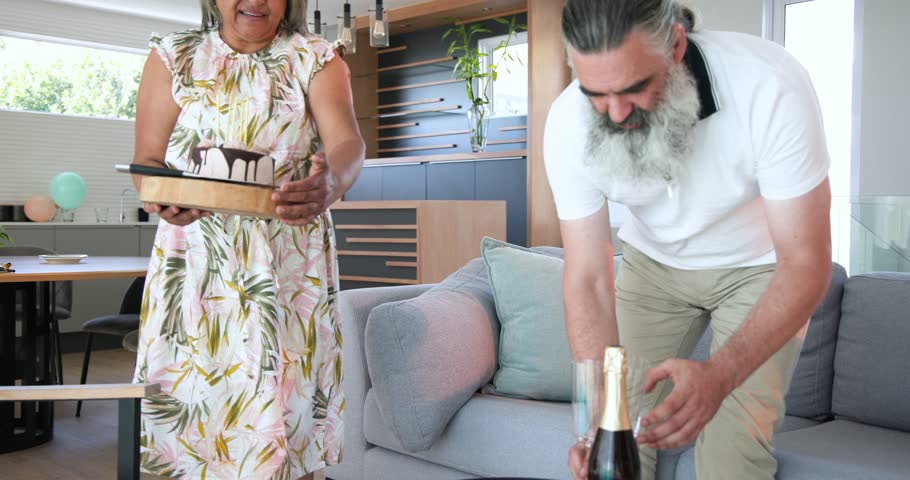 Senior couple carrying chocolate birthday cake toward coffee table placing candles in living room. Celebration, intimacy, elegance, companionship, home, joy, warmth