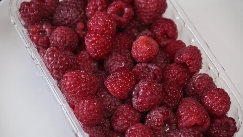 Close-up shot of fresh red raspberries in a transparent container. The camera slowly pans back, showcasing the ripe, juicy berries on a clean white background, ready for healthy eating.