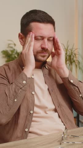Overworked young Caucasian businessman suffering from eye strain and headache working on laptop sitting at home office desk. Tired freelancer man guy takes off glasses and massages, rubbing temples.