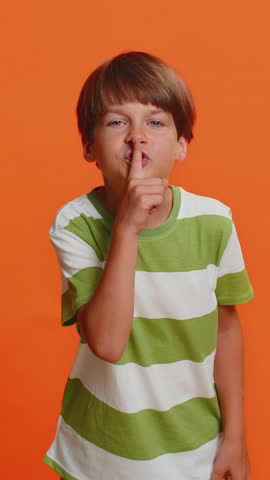 Shh be quiet please. Young preteen child boy kid presses index finger to lips makes silence gesture sign do not tells secret, stop talk gossip, confidential privacy isolated on orange background