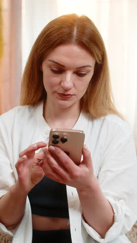 Middle-aged woman scrolls comments on smartphone at home, frowning and waving her hand with frustration stress. Young adult girl shows nervous reaction to negative feedback, visibly annoyed and tense.