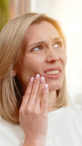 Middle-aged woman sits on home sofa holding cheek, wincing in visible discomfort from tooth pain. Adult ill lady expresses sharp ache with tense face and body language, looking for relief and calm.