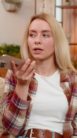Young adult woman sits on home sofa recording voice message on smartphone explaining with energy and emotion. Blonde girl gestures actively expressing herself clearly while reacting during voice input