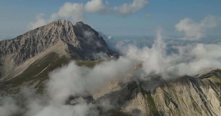 Aerial view of majestic mountains with rugged peaks and slopes, partially obscured by ethereal white clouds, contrasting with the clear blue sky, Gran Sasso, Abruzzo, Italy.