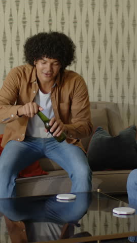 Vertical video: African American man twisting off beer cap, receiving bottle and toasting on sofa. Friendship, leisure, relaxation, celebration, socializing, casual, vertical video