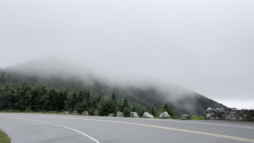 Dense fog slowly passing in front of a mountain, partially obscuring the peaks. Paved road runs through the foreground. Ideal for themes of travel, adventure, solitude, and nature