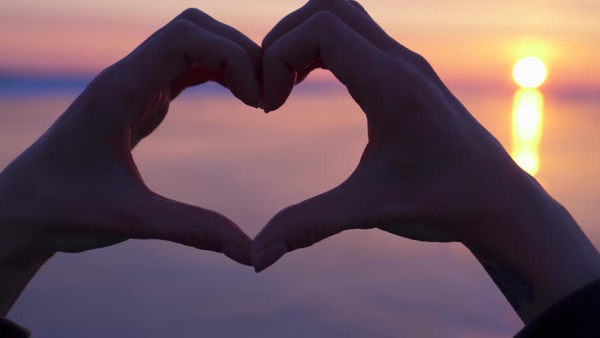 Hands forming heart shape against orange sunset over frozen lake. Symbol of love for nature and humanity, atmospheric abstract winter scene, majestic northern landscape backdrop. Traveler expresses