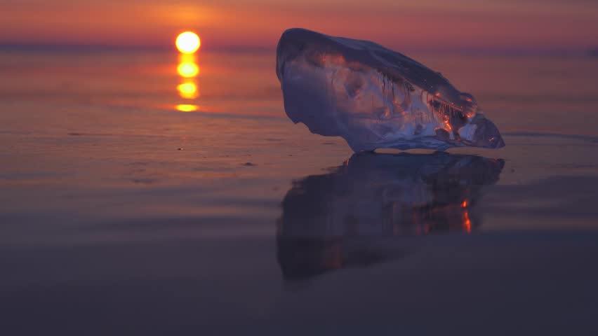 Cinematic sunset over Lake Baikal: crystal clear ice fragment rests on frozen surface. Siberian winter magic, sunlight refracting through orange ice, professional gimbak footage. Scenic details 4k
