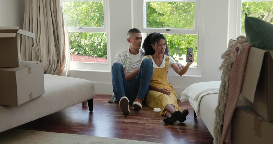 Diverse couple sitting on floor taking selfie while unpacking in new home, smiling happily. Relocation, moving, happiness, technology, lifestyle, bonding