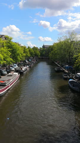 Beautifully picturesque Amsterdam canal scene that showcases charming boats gliding through vibrant greenery, all set under a clear blue skyperfect for inspiring travelthemed projects or artwork
