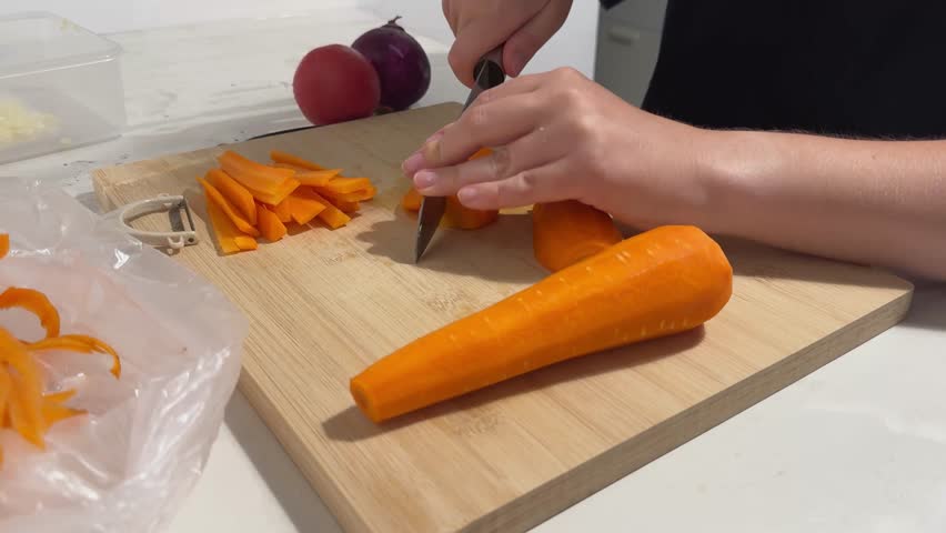Woman slices fresh orange carrot on light-colored wooden cutting board in indoor kitchen during the daytime