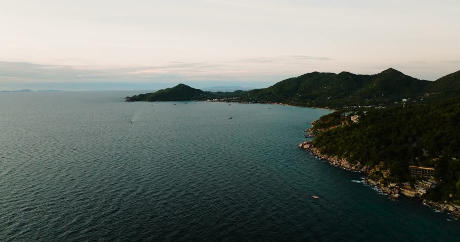 Scenic coastal view of lush green hills meeting calm waters with boats along the shoreline. Koh Tao, Thailand.