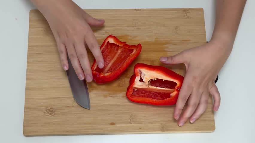 Woman uses a knife to remove seeds from a red pepper on a wooden cutting board at a kitchen table