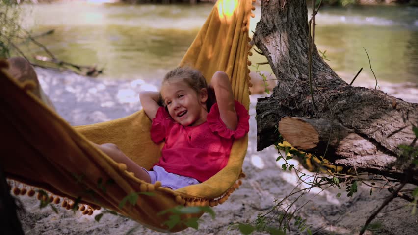 Carefree young girl swinging in lush riverside hammock, sharing joyful moments with mother amid serene natural landscape, laughing and playing together