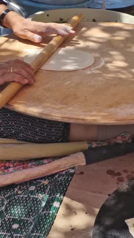 Hands of a woman rolling out unleavened bread for gözleme (a savoury Turkish pancake) on a round table while another turns gözleme being cooked on a wide, round sac griddle, at a local food festival