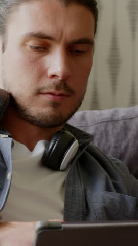 Vertical video: 30s man browsing tablet on couch checking smartwatch alert and adjusting headphones. Technology, relaxation, home, modern, connectivity, leisure, vertical video