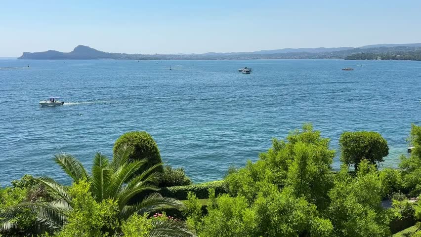 Scenic view from Isola del Garda overlooking Lake Garda in Italy. Clear blue waters, green trees, and distant mountains create a peaceful summer landscape with boats floating on the horizon