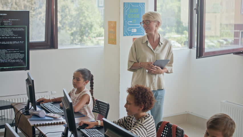 Young teacher of programming walking along row of diligent schoolkids while looking at their computer screens and checking individual assignment at lesson