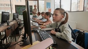Serious schoolgirl looking at computer screen and clicking mouse, then turning her head, listening to teacher and raising hand with the rest of class at lesson of programming - Powered by Shutterstock - Get 15% off with code: PIKWIZARD15