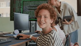 Smiling diligent schoolboy wearing striped pullover looking at camera while sitting by desk and keeping hand on computer mouse at lesson of programming against teacher helping girl - Powered by Shutterstock - Get 15% off with code: PIKWIZARD15