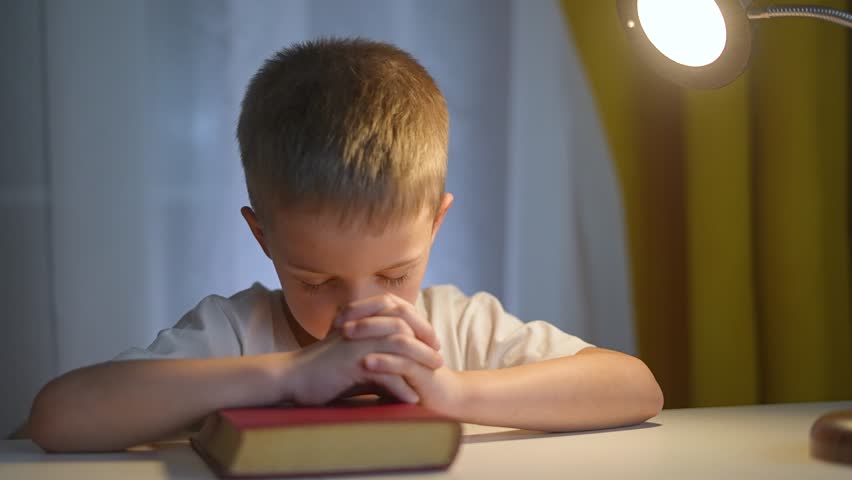 Boy sits alone in quiet room, praying in front old bible. Lamp beside boy creates gentle shadows as praying continues. Boy calm face and steady hands reflect worship devotion and peaceful hope faith