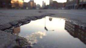 A close-up view of a puddle on an asphalt street, reflecting a blurred city skyline and sky during golden hour. Ripples disturb the calm surface, showing urban life mirrored in water. - Powered by Shutterstock - Get 15% off with code: PIKWIZARD15