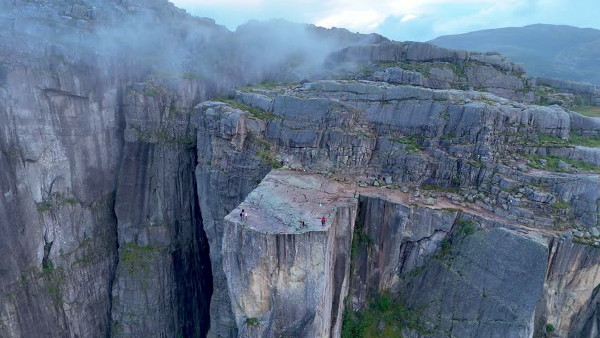 Experience the stunning beauty of Preikestolen in Norway, where daring adventurers stand on the iconic cliff, overlooking dramatic landscapes and rolling fog. Adventure awaits in this serene setting.