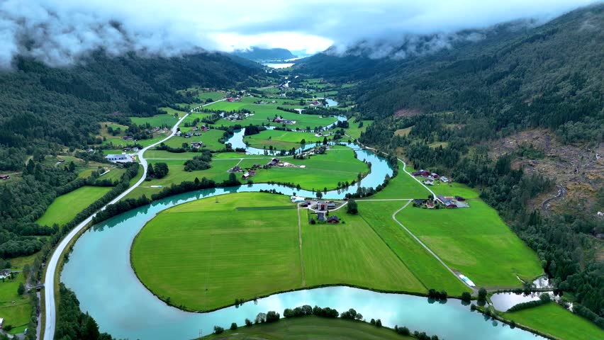 Breathtaking vista of Stryn, Norway showcasing vibrant green fields and meandering rivers. The serene landscape is framed by majestic mountains and shrouded in mist, creating a tranquil atmosphere.