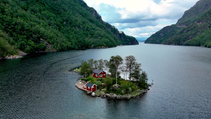 Nestled in a calm fjord, a vibrant red cabin sits on a small island embraced by stunning mountain landscapes and clear skies in Norway