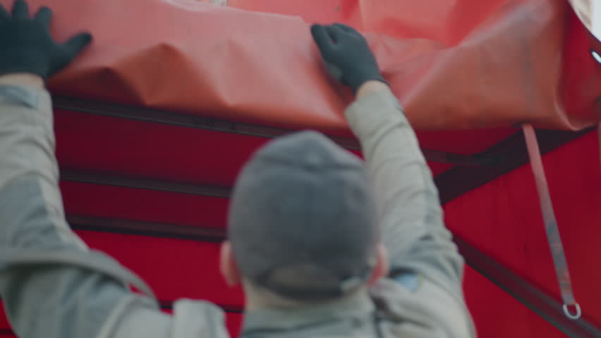 Back view of worker in uniform raising red truck tarp cover with both gloved hands during cargo preparation under daylight, revealing vehicle frame structure inside transport truck compartment