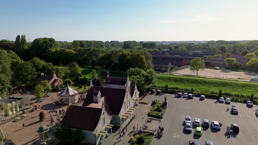 Utrecht, Utrecht region, Netherlands - 09.06.2025: Aerial view of a small amusement park with a carousel, colorful houses, parking lot, and greenery, located near a lake with sailing boats.