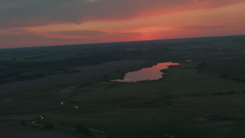 Scenic landscape featuring lake reflecting vibrant red orange dusk sky above expansive flat farmland under dramatic cloud canopy with glow and winding creek creating peaceful evening vista