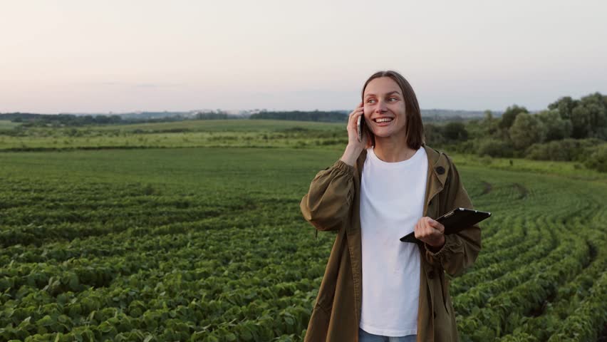 Modern agribusiness. Happy female farmer with digital tablet, talks on smartphone after examine and check in soybeans plants at field. Agronomist controls growth of harvest. Smart farming technology.
