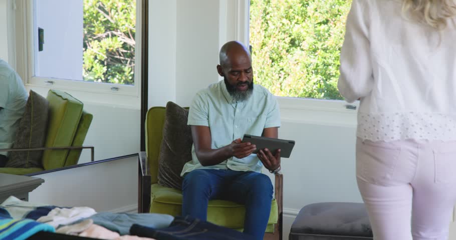 Diverse couple reviewing tablet at home as woman entering with keys, tapping, sorting clothes. Digital literacy, intergenerational, modern lifestyle, domestic scene, cozy environment, technology