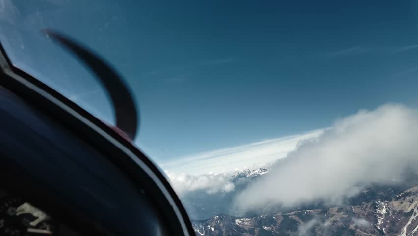 Scenic view through small aircraft propeller as plane flies through clouds over Alpine ridge and mountain peaks in Austria, under bright blue sky on a sunny day.
