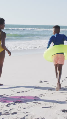 Vertical video: African American mom guiding kids off mat walking with neon and pink tubes to ocean. Family, beach, sunscreen, summer, outdoor, adventure, vertical video