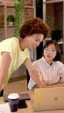 Vertical video: Diverse female colleagues at desk pointing at laptop trackpad explaining report. Collaboration, teamwork, modernoffice, communication, businesswomen, workplace, vertical video